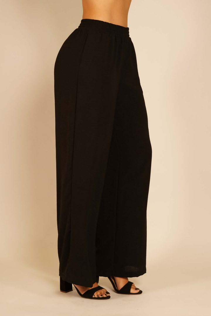 Pantalón Brena negro de seda Moma de pierna ancha con cintura alta elástica en modelo con tacones, vista lateral.