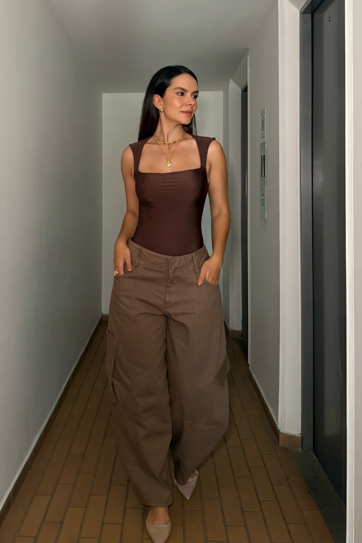 Modelo vistiendo Body Elsa Mocca control abdomen color marrón con pantalón ancho, caminando en un pasillo